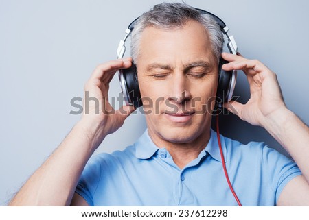 Relaxing music time. Portrait of senior man in headphones listening to music keeping eyes closed while standing against grey background