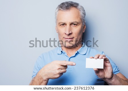 Call this number! Handsome senior man pointing on a business card and smiling while standing against grey background