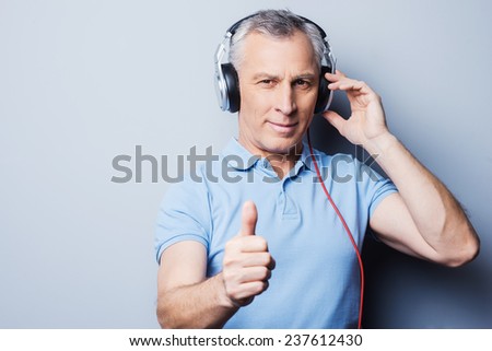 Listening to good music helps you at any situation. Portrait of senior man in headphones listening to music and showing his thumb up while standing against grey background