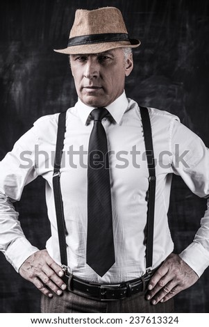 I am a boss! Confident senior man in hat and suspenders looking at camera and holding hands on hip while standing against dark background
