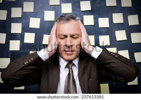 Tired from it all. Frustrated senior man in formalwear holding head in hands and keeping eyes closed while standing against blackboard with adhesive notes on it