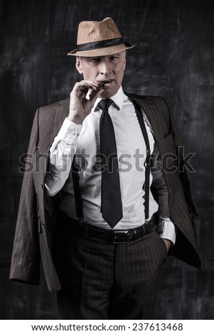 Powerful boss. Serious senior man in hat and suspenders smoking cigar and looking at you while standing against dark background