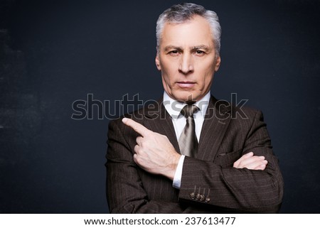 Pay attention to this. Portrait of confident senior man in formalwear looking at camera and pointing away while standing against blackboard