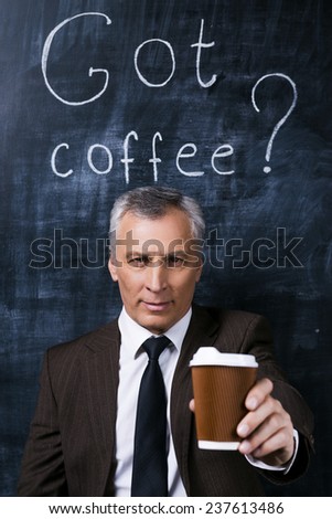 Take a break! Confident senior man in formalwear stretching out coffee cup and smiling while standing against blackboard with chalk drawing on it