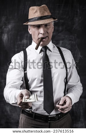 Money is not a problem. Serious senior man in hat and suspenders smoking cigar and stretching out money while standing against dark background