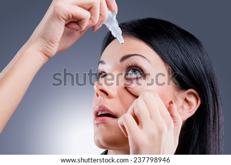 Taking care of her vision. Side view of young women applying eye drops while standing against grey background