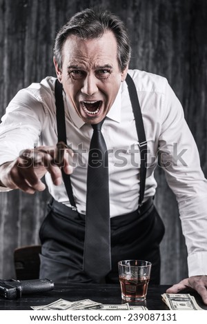 You are dead body! Furious senior man in shirt and suspenders shouting and pointing you while leaning at the table with lots of money laying near him