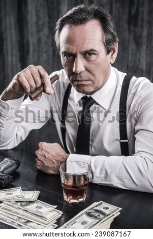 What do you want? Top view of serious senior man in shirt and suspenders sitting at the table and holding cigar while lots of money laying near him