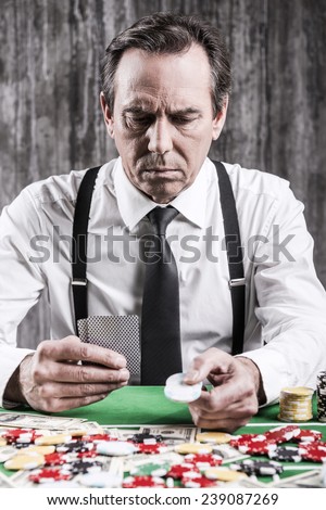 Confident poker player. Serious senior man in shirt and suspenders sitting at the poker table and holding cards and chips