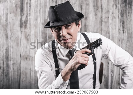 What will you say now? Bossy senior man in gangster clothing holding a gun and smiling while sitting at the chair against grey wall