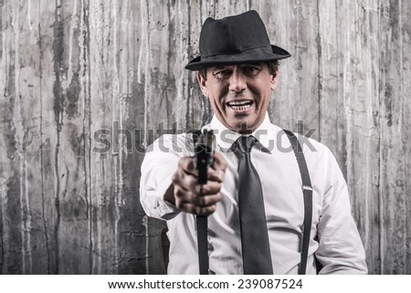 Say good bye! Bossy senior man in gangster clothing stretching out hand with gun while standing against grey wall