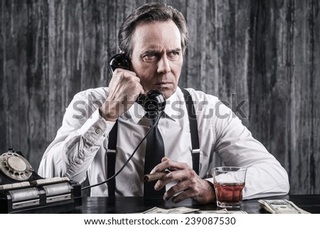 Tell me everything you know! Serious senior man in shirt and suspenders talking on the old-fashioned telephone while sitting at the table and smoking cigar