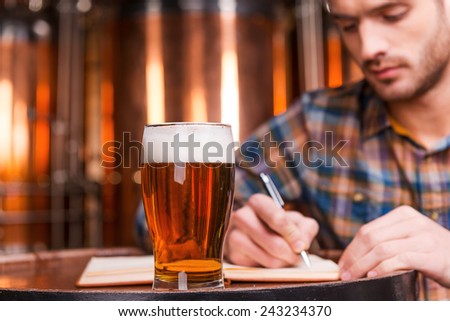 Planning his beer business. Confident young man in casual shirt writing something in his note pad while leaning at the wooden barrel with metal containers in the background