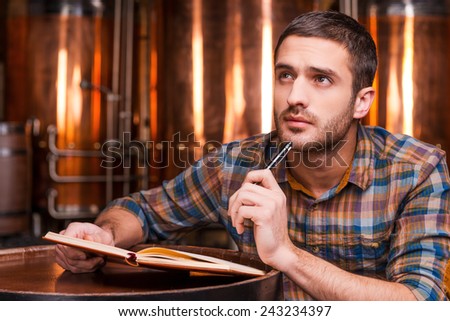 Generating ideas for his brewery. Thoughtful young man in casual shirt holding note pad and looking away while leaning at the wooden barrel with metal containers in the background