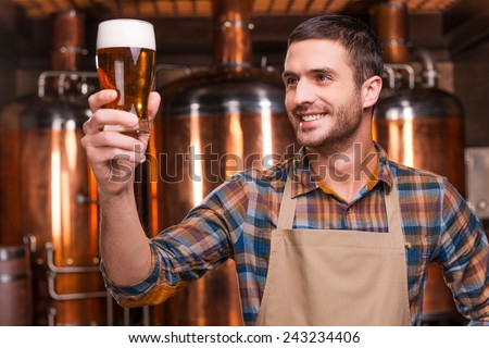 Happy brewer. Happy young male brewer in apron holding glass with beer and looking at it with smile while standing in front of metal containers