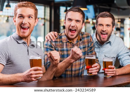 It is a goal! Three happy young men in casual wear holding glasses with beer and cheering while watching football match in bar together