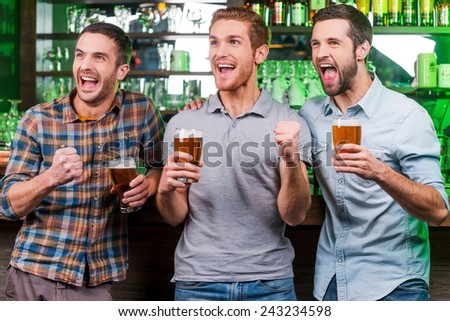 Goal! Three happy young men in casual wear holding glasses with beer and gesturing while standing at the bar counter and looking away