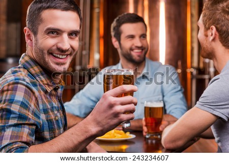 Spending time with friends. Handsome young man toasting with beer and smiling while sitting with his friends in beer pub