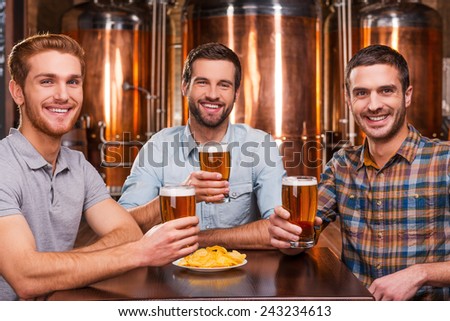 Friends in beer pub. Three cheerful young men in casual wear holding glasses with beer and smiling while sitting in beer pub and in front of metal containers