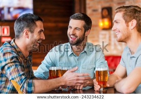 Meeting with the best friends. Three happy young men in casual wear talking and drinking beer while sitting in bar together