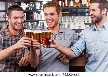 Cheers to us! Three happy young men in casual wear toasting with beer and smiling while standing at the bar counter together