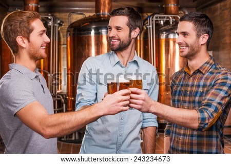 Cheers to us! Three cheerful young men in casual wear toasting with beer and smiling while standing in brewery in front of metal containers