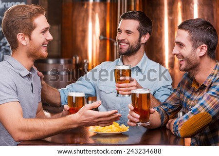 Spending great time with friends. Three happy young men in casual wear talking and drinking beer while sitting in beer pub together