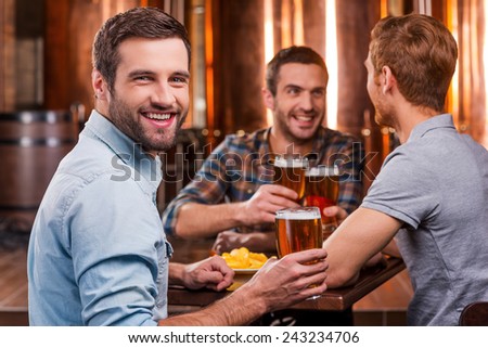 Spending time with best friends. Handsome young man toasting with beer and smiling while sitting with his friends in beer pub