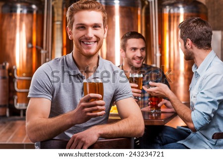 Spending time in beer pub. Handsome young man toasting with beer and smiling while his friends talking in the background