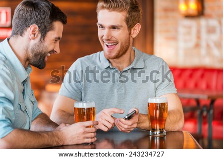 Just look at this photo! Two happy young men in casual wear drinking beer in pub while one of them holding smart phone and pointing it with smile