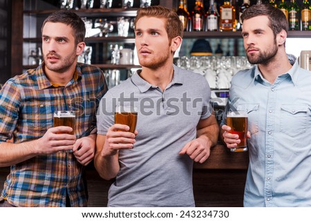 Watching football at the bar. Three handsome young men in casual wear holding glasses with beer and looking away while standing at the bar counter