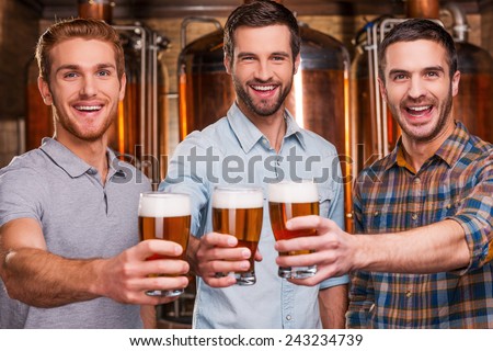 Cheers to you! Three cheerful young men in casual wear stretching out glasses with beer and smiling while standing in front of metal containers