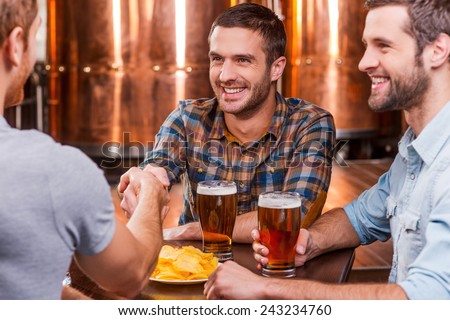 Meeting old friends. Three happy young men sitting in beer pub together while two of them handshaking