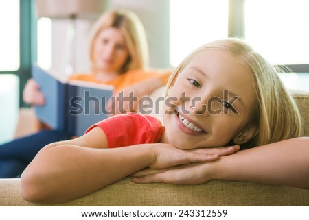 Enjoying time near mother. Happy little girl sitting on the couch and smiling while her mother reading book in the background