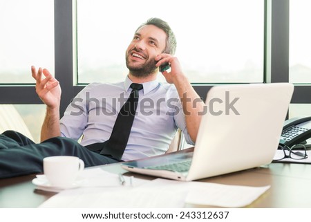 Good business talk. Handsome mature man in shirt and tie talking on the mobile phone and smiling while sitting at his working place