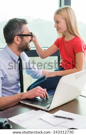 I need your glasses! Cheerful father in shirt and tie working on laptop while his playful daughter sitting close to him and touching his eyeglasses