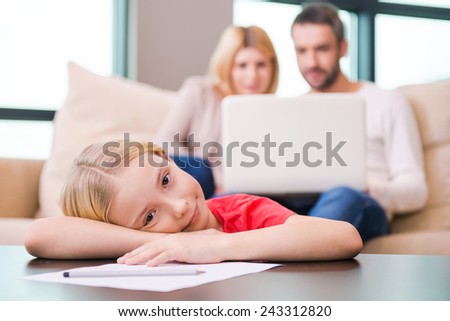 Leisure family time. Happy little girl leaning her face on table and smiling while her parents sitting in the background with laptop