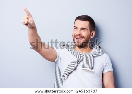 Look over there! Handsome young man pointing away and smiling while standing against grey background