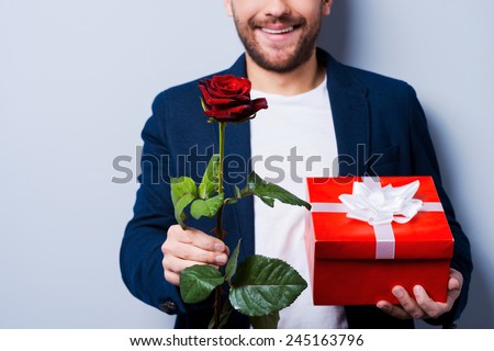 For sweetheart. Close-up of young man giving a gift and a flower to you while standing against grey background