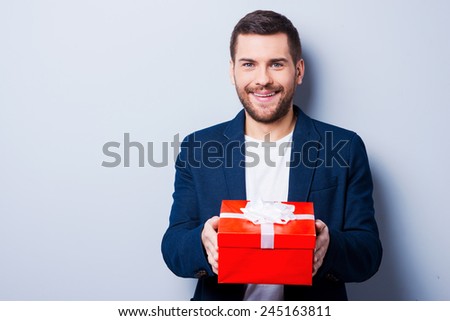 This gift is for you. Handsome young man in formalwear giving a gift to you while standing against grey background