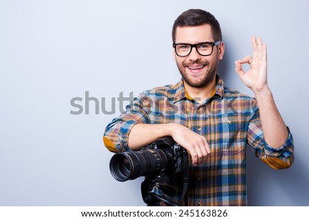 Love my job. Portrait of confident young man in shirt holding hand on camera on tripod and gesturing while standing against grey background