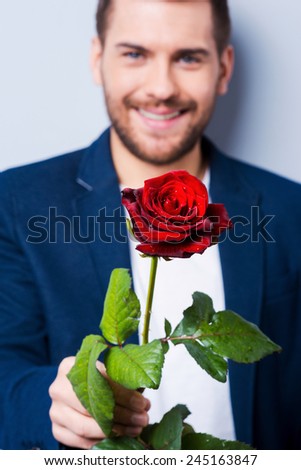 Rose for you. Handsome young man giving a flower to you while standing against grey background