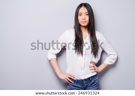Confident beauty. Beautiful young Asian woman looking at camera and holding hands on hips while standing against grey background