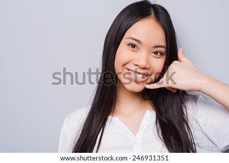Call me! Attractive young Asian woman looking at camera and gesturing while standing against grey background