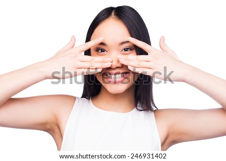 Playful beauty. Beautiful young Asian woman in pretty dress looking at camera and covering her face by hands while standing against white background