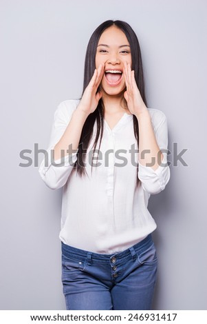 Telling the great news. Attractive young Asian woman shouting and holding hand near mouth while standing against grey background