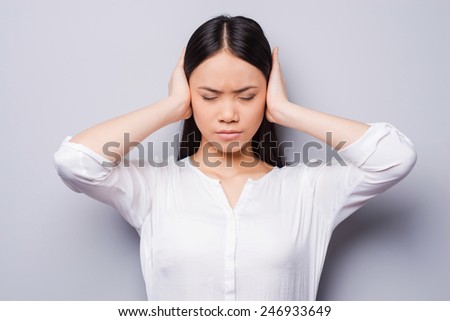 Too loud sound. Beautiful young Asian women covering ears with hands and keeping eyes closed while standing against grey background