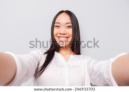 Happy moments must be saved. Cheerful young Asian woman holding camera and making selfie while standing against grey background