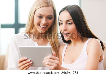 Can you believe it?! Two beautiful young women looking at the digital tablet and looking surprised while both sitting on the couch together