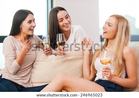 Discussing the latest gossips. Three beautiful young women talking and smiling while drinking wine and sitting on the couch together
